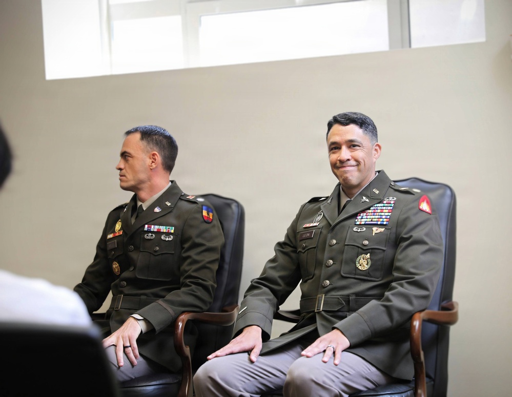 Two U.S. Army officers in Army Green Service Uniforms (AGSU) are seated in leather chairs, appearing to be attending a formal military event or ceremony. The officer on the right displays an extensive array of ribbons, badges, and decorations indicating senior rank and significant service history. Both officers appear relaxed, with the right officer smiling slightly toward an unseen audience or presenter.