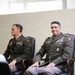 Two U.S. Army officers in Army Green Service Uniforms (AGSU) are seated in leather chairs, appearing to be attending a formal military event or ceremony. The officer on the right displays an extensive array of ribbons, badges, and decorations indicating senior rank and significant service history. Both officers appear relaxed, with the right officer smiling slightly toward an unseen audience or presenter.