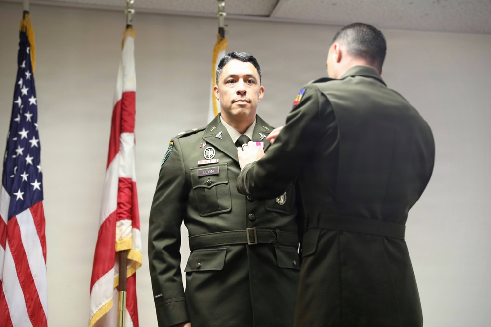A military award ceremony is taking place indoors, where one uniformed soldier is pinning a medal or decoration onto another soldier's chest. The recipient, whose name tag reads 'LEON,' stands at attention in Army dress green uniform while facing the camera. An American flag and what appears to be a state flag are visible in the background.