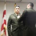 A military award ceremony is taking place indoors, where one uniformed soldier is pinning a medal or decoration onto another soldier's chest. The recipient, whose name tag reads 'LEON,' stands at attention in Army dress green uniform while facing the camera. An American flag and what appears to be a state flag are visible in the background.