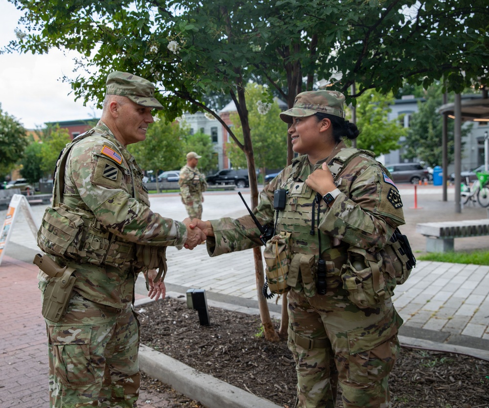 Two U.S. Army soldiers in Operational Camouflage Pattern (OCP) uniforms are shaking hands in an urban outdoor setting. The soldier on the left displays a 3rd Infantry Division patch and appears to be of higher rank, while the soldier on the right is wearing body armor and carrying a radio. A third soldier is visible in the background near what appears to be a public plaza or street.