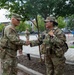 Two U.S. Army soldiers in Operational Camouflage Pattern (OCP) uniforms are shaking hands in an urban outdoor setting. The soldier on the left displays a 3rd Infantry Division patch and appears to be of higher rank, while the soldier on the right is wearing body armor and carrying a radio. A third soldier is visible in the background near what appears to be a public plaza or street.