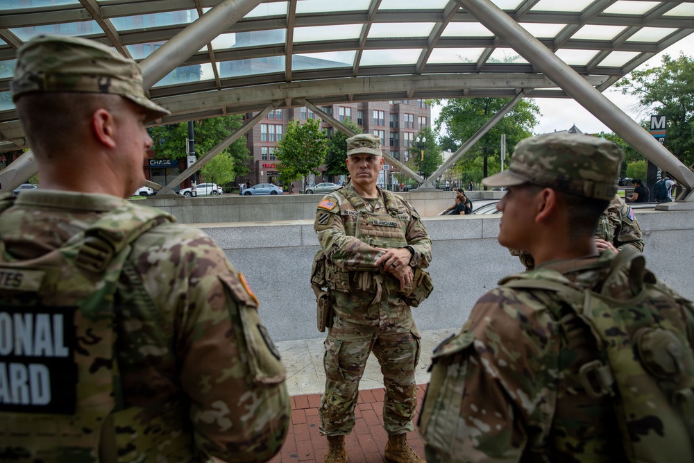 Three U.S. National Guard soldiers in multicam/OCP camouflage uniforms are gathered near what appears to be a Metro station entrance, identifiable by the distinctive 'M' sign in the background. The soldiers appear to be in a briefing or conversation, with one soldier facing the other two. The setting appears to be an urban area with commercial buildings, trees, and parked vehicles visible in the background.
