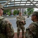 Three U.S. National Guard soldiers in multicam/OCP camouflage uniforms are gathered near what appears to be a Metro station entrance, identifiable by the distinctive 'M' sign in the background. The soldiers appear to be in a briefing or conversation, with one soldier facing the other two. The setting appears to be an urban area with commercial buildings, trees, and parked vehicles visible in the background.