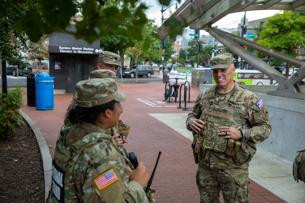 Three U.S. Army soldiers in OCP (Operational Camouflage Pattern) uniforms are conversing near the Eastern Market Metro Station in Washington, D.C. The soldier on the right, identified by name tape as 'BLANCHARD,' appears to be a senior officer engaging with two other soldiers, one of whom is holding a radio. The setting suggests a military presence or patrol operation in an urban environment.