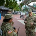 Three U.S. Army soldiers in OCP (Operational Camouflage Pattern) uniforms are conversing near the Eastern Market Metro Station in Washington, D.C. The soldier on the right, identified by name tape as 'BLANCHARD,' appears to be a senior officer engaging with two other soldiers, one of whom is holding a radio. The setting suggests a military presence or patrol operation in an urban environment.