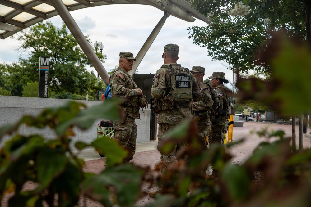 A group of National Guard soldiers in camouflage uniforms are gathered near the Eastern Market Metro station in Washington, D.C. At least three to four personnel are visible, with one soldier's vest clearly labeled 'NATIONAL GUARD' on the back. The scene appears to be a routine assembly or briefing near a public transit entrance.