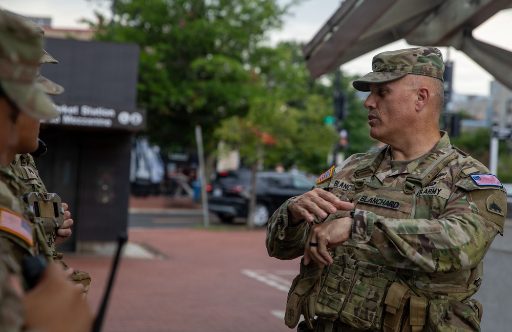 A U.S. Army soldier named Blanchard is engaged in conversation with another soldier in an urban setting. Both personnel are wearing OCP (Operational Camouflage Pattern) uniforms with tactical vests and American flag patches. The background shows what appears to be a metro or transit station entrance in an urban area.