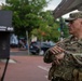 A U.S. Army soldier named Blanchard is engaged in conversation with another soldier in an urban setting. Both personnel are wearing OCP (Operational Camouflage Pattern) uniforms with tactical vests and American flag patches. The background shows what appears to be a metro or transit station entrance in an urban area.