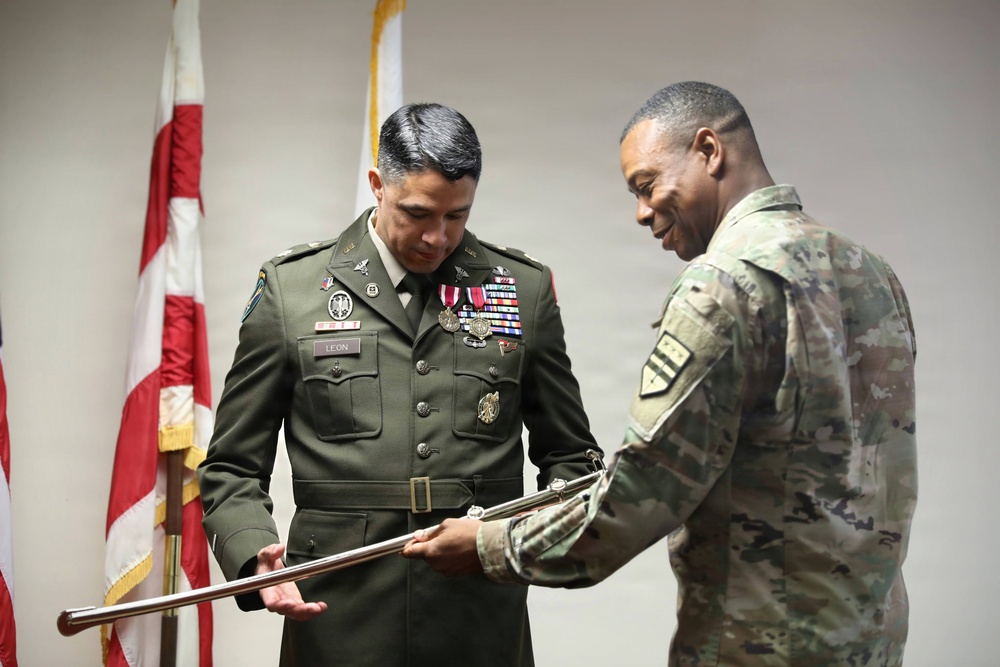 Two U.S. Army soldiers are participating in a formal military ceremony, with one soldier in dress greens (Class A uniform) and the other in Army Combat Uniform (ACU/OCP). They are jointly holding what appears to be a ceremonial saber or sword, suggesting a retirement, promotion, or change-of-command ceremony. An American flag and what appears to be a unit guidon are visible in the background.