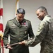 Two U.S. Army soldiers are participating in a formal military ceremony, with one soldier in dress greens (Class A uniform) and the other in Army Combat Uniform (ACU/OCP). They are jointly holding what appears to be a ceremonial saber or sword, suggesting a retirement, promotion, or change-of-command ceremony. An American flag and what appears to be a unit guidon are visible in the background.