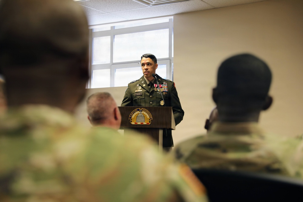 A military officer in dress green uniform is speaking at a podium bearing a 'Capital Guardian' insignia, addressing an audience of soldiers in camouflage uniforms. The setting appears to be an indoor military briefing room or ceremony hall. The speaker is wearing multiple medals and ribbons, suggesting a formal military ceremony or award presentation.