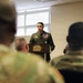 A military officer in dress green uniform is speaking at a podium bearing a 'Capital Guardian' insignia, addressing an audience of soldiers in camouflage uniforms. The setting appears to be an indoor military briefing room or ceremony hall. The speaker is wearing multiple medals and ribbons, suggesting a formal military ceremony or award presentation.