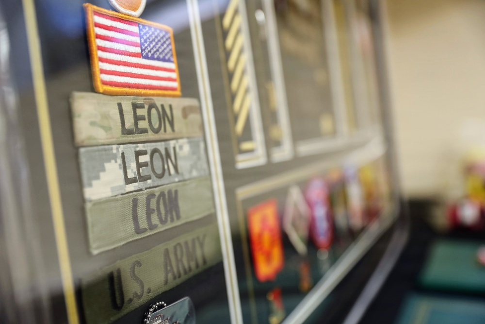A military shadow box or display case containing U.S. Army memorabilia belonging to a service member with the surname 'LEON'. The display includes multiple name tapes in different camouflage patterns, a U.S. flag patch, and various military insignia and ribbons. The items represent a collection of uniform elements and service memorabilia typical of a military career display.