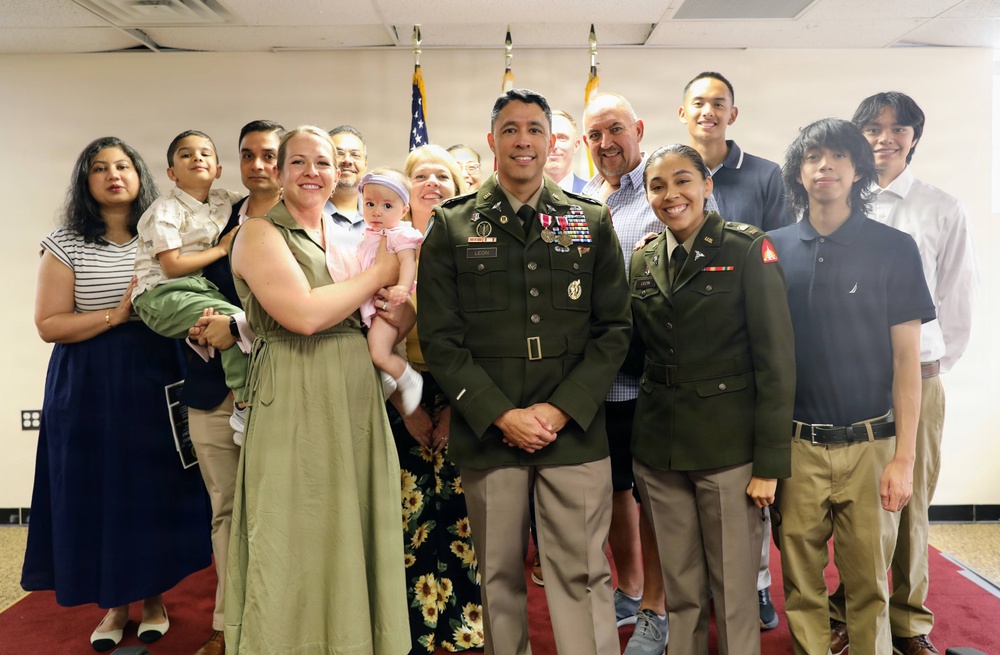 A formal military ceremony photograph showing two U.S. Army officers in dress green uniforms standing at the center, surrounded by approximately 14 family members and guests. The setting appears to be an indoor military facility with American flags visible in the background. The occasion appears to be a promotion or award ceremony based on the medals and insignia visible on the uniforms.
