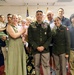 A formal military ceremony photograph showing two U.S. Army officers in dress green uniforms standing at the center, surrounded by approximately 14 family members and guests. The setting appears to be an indoor military facility with American flags visible in the background. The occasion appears to be a promotion or award ceremony based on the medals and insignia visible on the uniforms.
