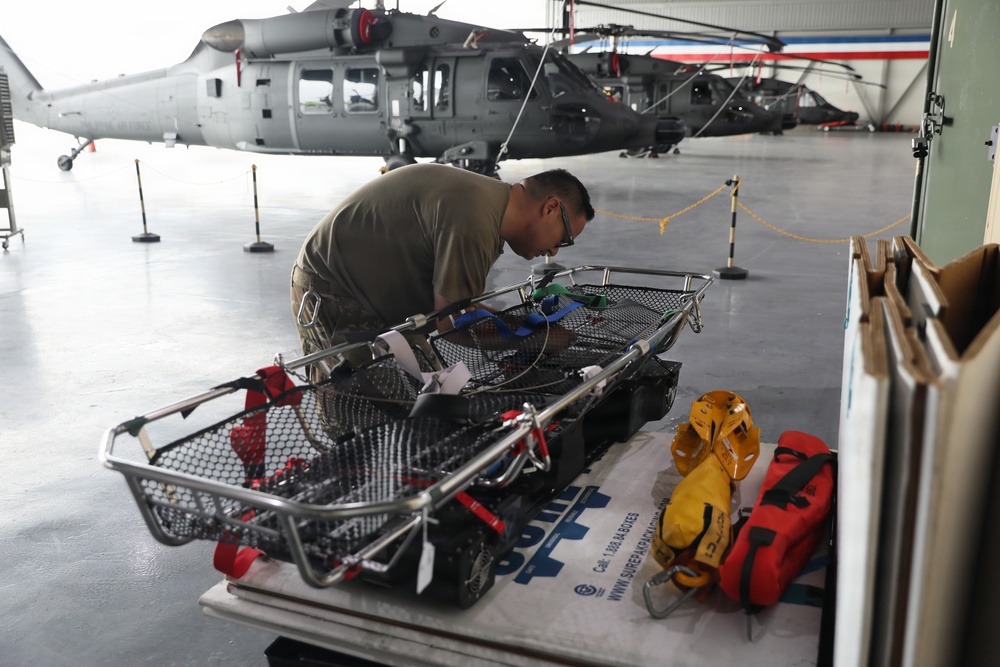 A military or rescue personnel in olive/tan uniform is inspecting or preparing a Stokes litter (rescue basket) in a helicopter hangar. In the background, at least two gray military helicopters are visible, consistent with UH-60 Black Hawk variants. The scene suggests pre-mission equipment preparation for search and rescue operations.
