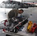 A military or rescue personnel in olive/tan uniform is inspecting or preparing a Stokes litter (rescue basket) in a helicopter hangar. In the background, at least two gray military helicopters are visible, consistent with UH-60 Black Hawk variants. The scene suggests pre-mission equipment preparation for search and rescue operations.