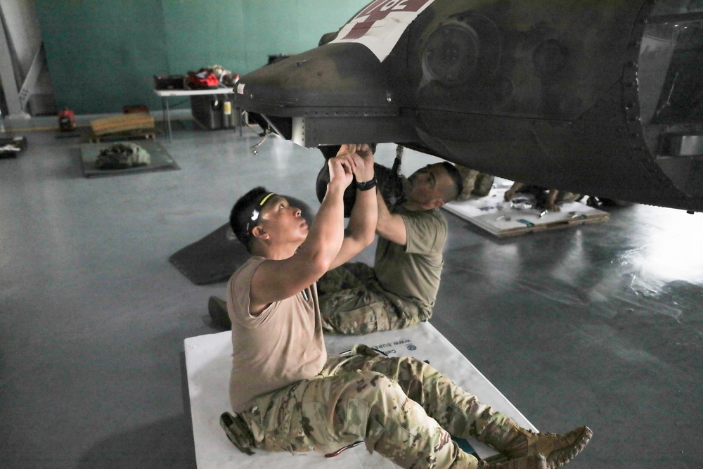 Two military personnel in camouflage uniforms are performing maintenance work on the underside of a military helicopter inside a hangar. They are lying/sitting on protective mats on the floor while working on components beneath the aircraft. The setting appears to be an active maintenance bay with tools and equipment visible in the background.