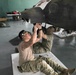 Two military personnel in camouflage uniforms are performing maintenance work on the underside of a military helicopter inside a hangar. They are lying/sitting on protective mats on the floor while working on components beneath the aircraft. The setting appears to be an active maintenance bay with tools and equipment visible in the background.