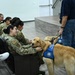 A therapy or facility dog wearing a blue service vest is being introduced to military personnel seated in an auditorium or briefing room. A civilian handler holds the golden retriever on a leash while a smiling service member in camouflage uniform pets the dog. The setting appears to be a morale or wellness event on a military installation.