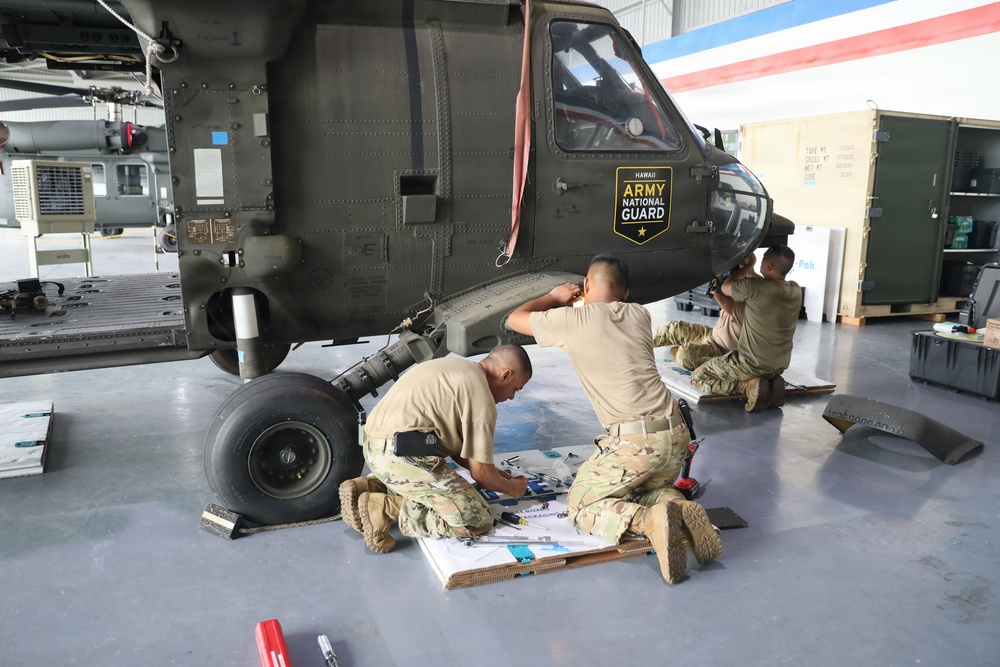 Three soldiers in camouflage uniforms are performing maintenance work on a military helicopter inside a hangar. The helicopter bears the 'Hawaii Army National Guard' insignia on its fuselage. Two soldiers are kneeling on the floor working with parts and documents, while a third works on the side of the aircraft.