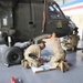 Three soldiers in camouflage uniforms are performing maintenance work on a military helicopter inside a hangar. The helicopter bears the 'Hawaii Army National Guard' insignia on its fuselage. Two soldiers are kneeling on the floor working with parts and documents, while a third works on the side of the aircraft.