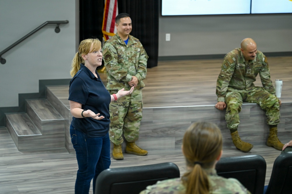A civilian woman in a navy blue polo shirt and jeans appears to be presenting or speaking to a group in a modern briefing or meeting room. Two military personnel in OCP (Operational Camouflage Pattern) uniforms are visible in the background, one standing and one seated on a raised platform. An American flag and a projection screen are visible in the background.