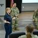 A civilian woman in a navy blue polo shirt and jeans appears to be presenting or speaking to a group in a modern briefing or meeting room. Two military personnel in OCP (Operational Camouflage Pattern) uniforms are visible in the background, one standing and one seated on a raised platform. An American flag and a projection screen are visible in the background.