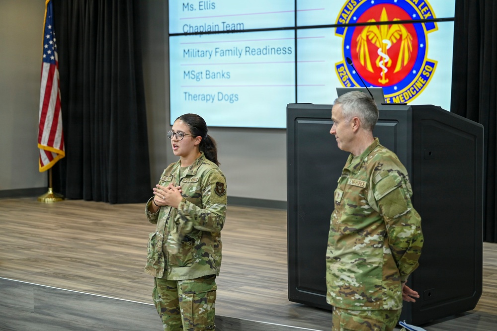 Two U.S. Air Force personnel in OCP (Operational Camouflage Pattern) uniforms are presenting at a formal military briefing or event. A female airman is speaking while a senior male officer stands near a podium. A large display screen behind them shows an agenda and a military unit emblem identified as a Medical Squadron.