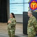 Two U.S. Air Force personnel in OCP (Operational Camouflage Pattern) uniforms are presenting at a formal military briefing or event. A female airman is speaking while a senior male officer stands near a podium. A large display screen behind them shows an agenda and a military unit emblem identified as a Medical Squadron.