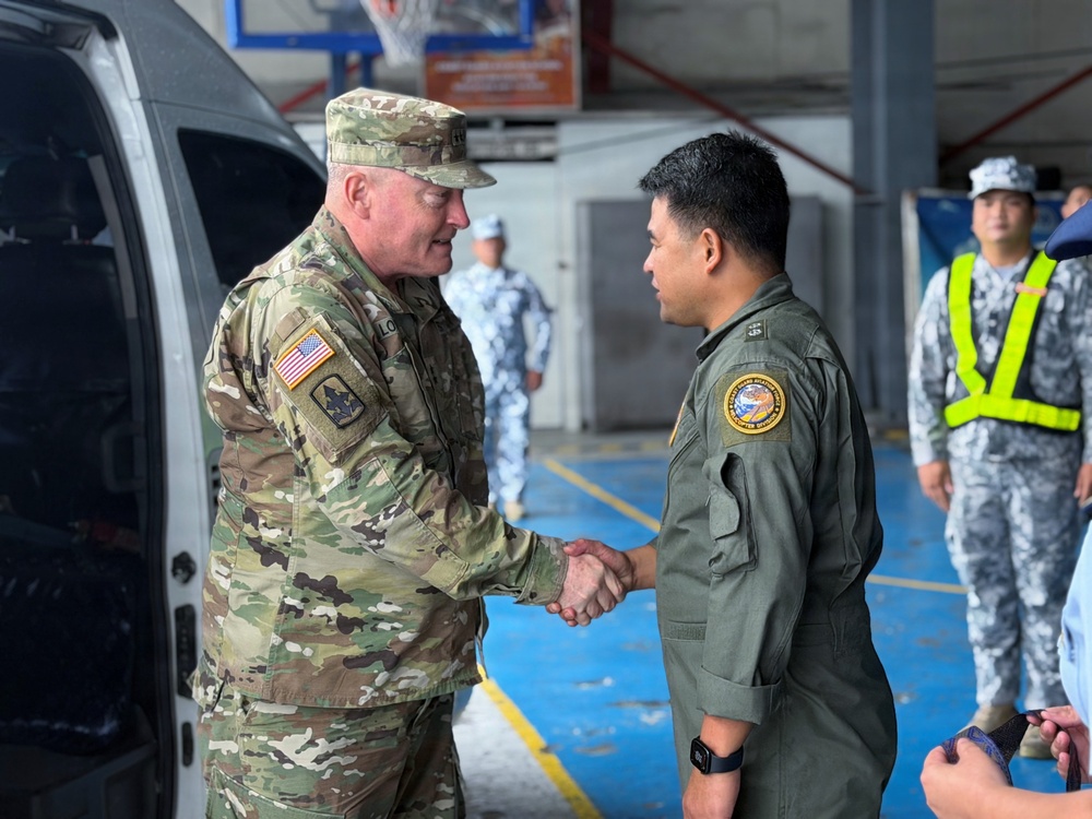 Two military officers are shaking hands in what appears to be a hangar or indoor military facility. A U.S. Army soldier in MultiCam/OCP camouflage uniform with an American flag patch is greeting a foreign military officer in an olive drab flight suit bearing a circular unit patch. Several other military personnel in light blue uniforms are visible in the background.