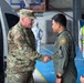 Two military officers are shaking hands in what appears to be a hangar or indoor military facility. A U.S. Army soldier in MultiCam/OCP camouflage uniform with an American flag patch is greeting a foreign military officer in an olive drab flight suit bearing a circular unit patch. Several other military personnel in light blue uniforms are visible in the background.