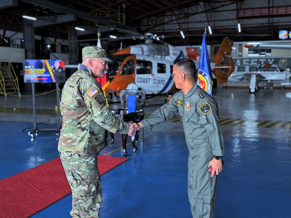 Two military officers are shaking hands inside an aviation hangar, with a Coast Guard helicopter visible in the background. The officer on the left wears US Army multicam (OCP) uniform with an American flag patch, while the officer on the right wears a flight suit with what appears to be Philippine military insignia. A welcome banner and organizational flags are visible, suggesting a formal reception or bilateral meeting.