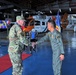 Two military officers are shaking hands inside an aviation hangar, with a Coast Guard helicopter visible in the background. The officer on the left wears US Army multicam (OCP) uniform with an American flag patch, while the officer on the right wears a flight suit with what appears to be Philippine military insignia. A welcome banner and organizational flags are visible, suggesting a formal reception or bilateral meeting.