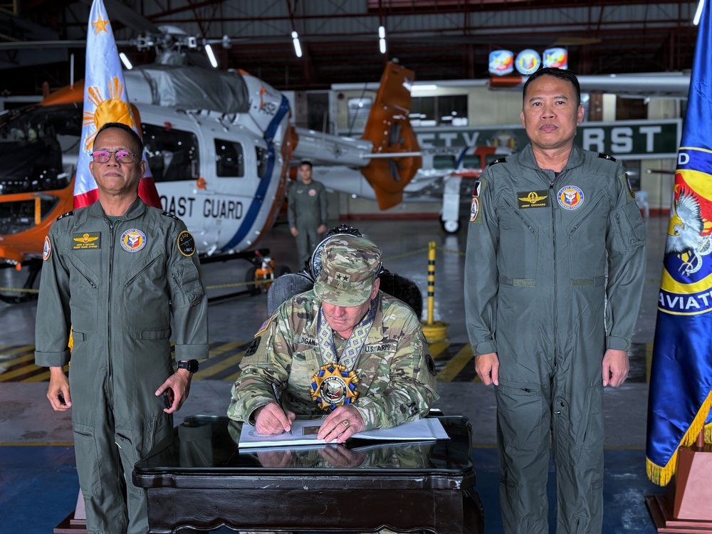 A formal military signing ceremony taking place inside an aviation hangar, featuring a U.S. Army officer seated at a desk signing documents, flanked by two Philippine military officers in flight suits. A Philippine Coast Guard helicopter is visible in the background along with Philippine and military unit flags. The setting suggests a bilateral military cooperation or agreement signing event.