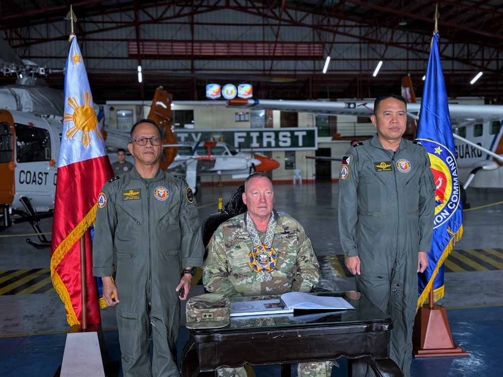 Three military personnel are posed in an aviation hangar, with a U.S. Army officer seated at a desk flanked by two Philippine Coast Guard officers in flight suits. The setting appears to be an official ceremony or signing event, with the Philippine national flag and a Philippine Coast Guard Aviation Command flag displayed on either side. Aircraft and Coast Guard equipment are visible in the background.