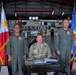 Three military personnel are posed in an aviation hangar, with a U.S. Army officer seated at a desk flanked by two Philippine Coast Guard officers in flight suits. The setting appears to be an official ceremony or signing event, with the Philippine national flag and a Philippine Coast Guard Aviation Command flag displayed on either side. Aircraft and Coast Guard equipment are visible in the background.