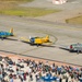 An aerial view of an airshow featuring three vintage World War II-era trainer aircraft taxiing on a runway in formation. The aircraft include a blue plane marked 'L99', a yellow plane marked 'RCAF 421', and a silver/gray plane with US Army Air Forces markings. A large crowd of spectators watches from a designated viewing area along the flight line, with Navy recruitment tents visible.