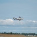 A vintage WWII-era training aircraft, likely a North American T-6 Texan, is captured in flight against a partly cloudy blue sky. The silver aircraft displays US military markings including a white star insignia on the fuselage and what appears to be a red spinner on the propeller. Below, a treeline and what appears to be an airfield or industrial facility are visible on the horizon.