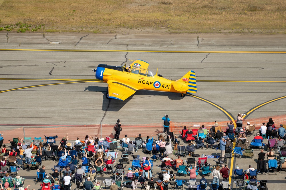 An aerial photograph captures a vintage yellow RCAF (Royal Canadian Air Force) training aircraft marked '421' taxiing on a runway at an airshow. The aircraft, identifiable as a North American Harvard/T-6 Texan trainer, is painted in classic RCAF yellow with blue accents and displays the Canadian maple leaf roundel. A large crowd of spectators seated in lawn chairs lines the tarmac below, watching and photographing the aircraft.