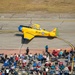 An aerial photograph captures a vintage yellow RCAF (Royal Canadian Air Force) training aircraft marked '421' taxiing on a runway at an airshow. The aircraft, identifiable as a North American Harvard/T-6 Texan trainer, is painted in classic RCAF yellow with blue accents and displays the Canadian maple leaf roundel. A large crowd of spectators seated in lawn chairs lines the tarmac below, watching and photographing the aircraft.