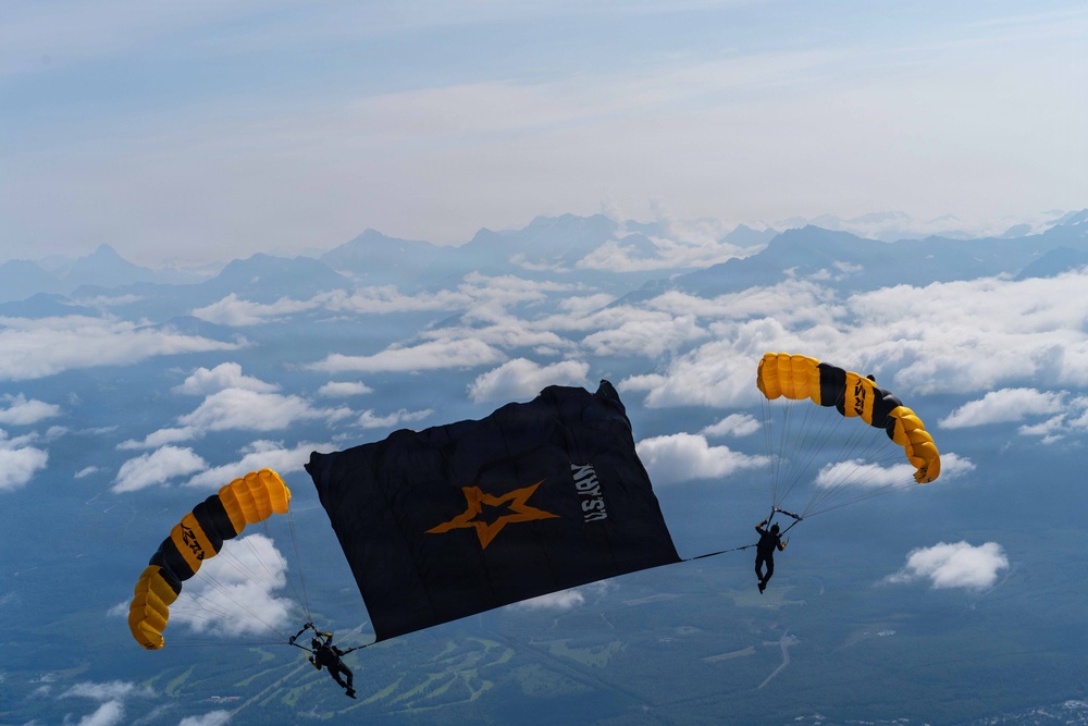 Two skydivers with matching black-and-gold parachutes are carrying a large black U.S. Army banner between them at high altitude. The jumpers appear to be members of the U.S. Army Golden Knights parachute demonstration team, identifiable by their distinctive gold and black canopies. A dramatic mountain range and cloud layer are visible in the background below them.