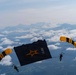 Two skydivers with matching black-and-gold parachutes are carrying a large black U.S. Army banner between them at high altitude. The jumpers appear to be members of the U.S. Army Golden Knights parachute demonstration team, identifiable by their distinctive gold and black canopies. A dramatic mountain range and cloud layer are visible in the background below them.
