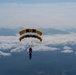 Two skydivers are performing a tandem parachute jump using a distinctive black and gold dual-canopy parachute system. They are descending above a cloud layer with a mountainous landscape visible in the background. The parachute design and color scheme suggest this may be a military or professional demonstration team.