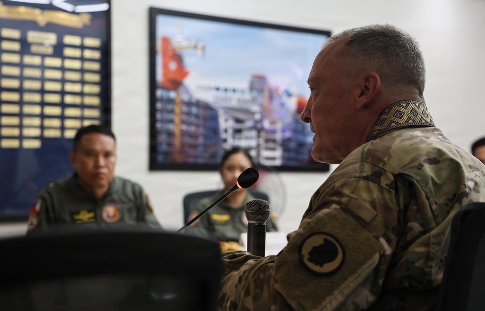 A military meeting or briefing is taking place between personnel from what appears to be two different nations. A senior officer in US Army multicam uniform with a decorative collar piece speaks into a microphone, while personnel in olive green flight suits with Philippine Air Force patches listen across the table. The setting appears to be an official military conference room with a display screen and honor board visible in the background.