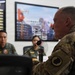 A military meeting or briefing is taking place between personnel from what appears to be two different nations. A senior officer in US Army multicam uniform with a decorative collar piece speaks into a microphone, while personnel in olive green flight suits with Philippine Air Force patches listen across the table. The setting appears to be an official military conference room with a display screen and honor board visible in the background.