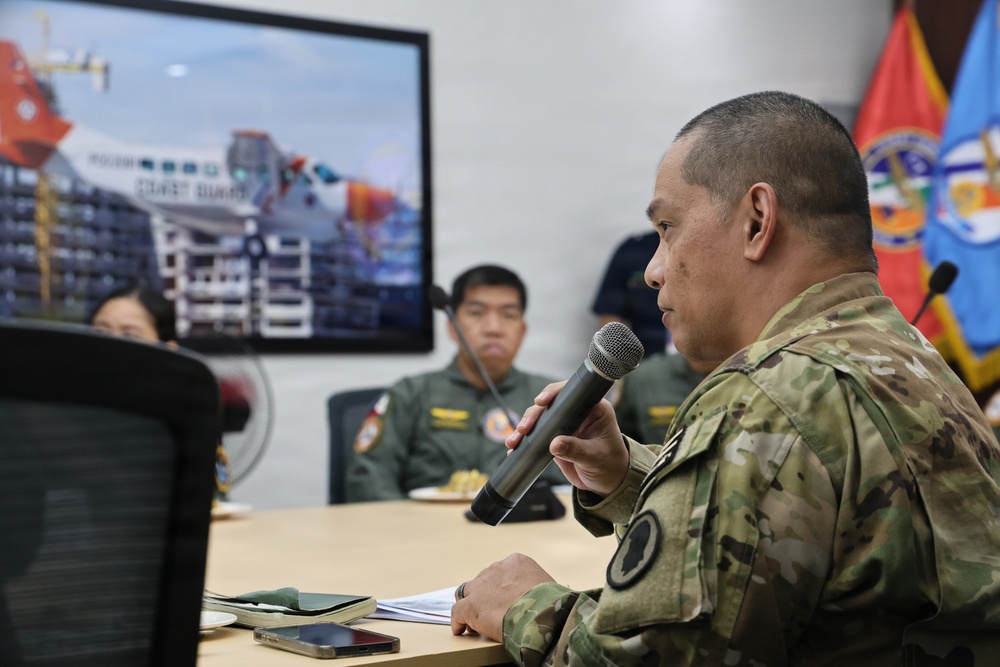 A military briefing or meeting is taking place in a conference room setting. A soldier in MultiCam/OCP camouflage uniform is speaking into a handheld microphone while seated at a table. Other military personnel in olive/flight suits are visible in the background, along with a display screen showing what appears to be a Coast Guard-related presentation.