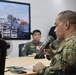 A military briefing or meeting is taking place in a conference room setting. A soldier in MultiCam/OCP camouflage uniform is speaking into a handheld microphone while seated at a table. Other military personnel in olive/flight suits are visible in the background, along with a display screen showing what appears to be a Coast Guard-related presentation.
