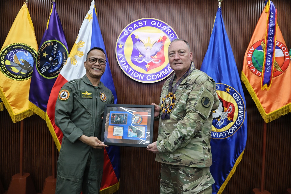 Two military officers are posing together in front of a Coast Guard Aviation Command seal and multiple organizational flags. They are jointly holding a framed commemorative plaque or shadow box, suggesting an official exchange or ceremony. One officer is wearing a Philippine Coast Guard flight suit and the other is in U.S. Army multicam (OCP) uniform.